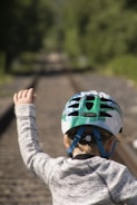 A child wearing a green and white helmet and a gray sweater is standing on train tracks, with one arm raised. The background features blurred greenery.
