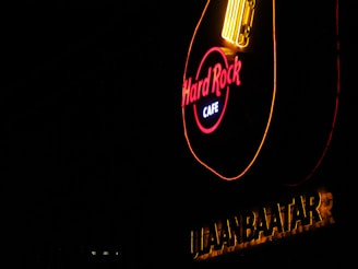 A brightly illuminated neon sign in the shape of a guitar featuring the words 'Hard Rock Cafe.' The sign is set against a dark background, with the word 'ULAANBAATAR' in bold letters underneath.