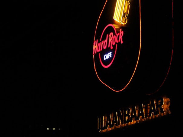 A brightly illuminated neon sign in the shape of a guitar featuring the words 'Hard Rock Cafe.' The sign is set against a dark background, with the word 'ULAANBAATAR' in bold letters underneath.