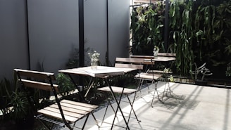 The image depicts an outdoor seating area with several wooden chairs and tables. The space is accentuated with a vertical garden featuring various green plants along the back wall, providing a natural and inviting atmosphere. A few small vases with flowers are placed on the tables, adding a touch of elegance. Shadows cast by the chairs and plants suggest sunlight, enhancing the serene setting.