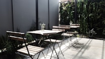 The image depicts an outdoor seating area with several wooden chairs and tables. The space is accentuated with a vertical garden featuring various green plants along the back wall, providing a natural and inviting atmosphere. A few small vases with flowers are placed on the tables, adding a touch of elegance. Shadows cast by the chairs and plants suggest sunlight, enhancing the serene setting.