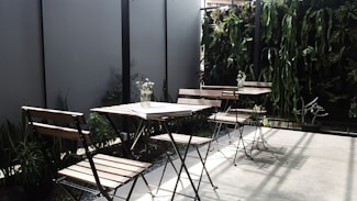 The image depicts an outdoor seating area with several wooden chairs and tables. The space is accentuated with a vertical garden featuring various green plants along the back wall, providing a natural and inviting atmosphere. A few small vases with flowers are placed on the tables, adding a touch of elegance. Shadows cast by the chairs and plants suggest sunlight, enhancing the serene setting.
