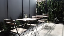 The image depicts an outdoor seating area with several wooden chairs and tables. The space is accentuated with a vertical garden featuring various green plants along the back wall, providing a natural and inviting atmosphere. A few small vases with flowers are placed on the tables, adding a touch of elegance. Shadows cast by the chairs and plants suggest sunlight, enhancing the serene setting.