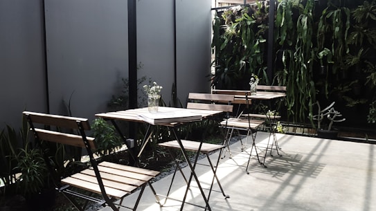 The image depicts an outdoor seating area with several wooden chairs and tables. The space is accentuated with a vertical garden featuring various green plants along the back wall, providing a natural and inviting atmosphere. A few small vases with flowers are placed on the tables, adding a touch of elegance. Shadows cast by the chairs and plants suggest sunlight, enhancing the serene setting.