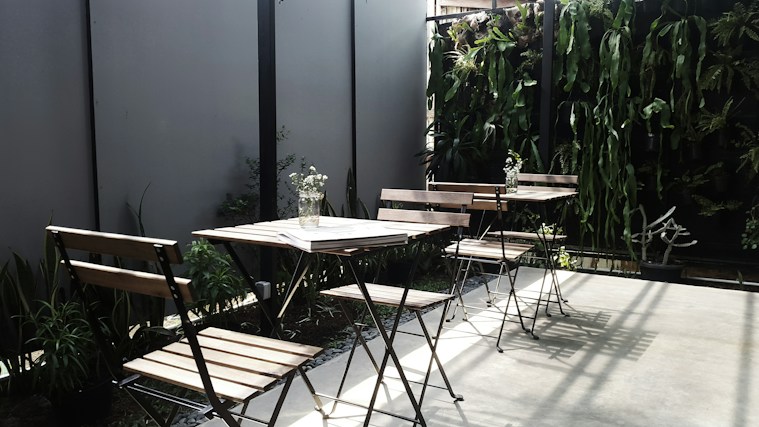 The image depicts an outdoor seating area with several wooden chairs and tables. The space is accentuated with a vertical garden featuring various green plants along the back wall, providing a natural and inviting atmosphere. A few small vases with flowers are placed on the tables, adding a touch of elegance. Shadows cast by the chairs and plants suggest sunlight, enhancing the serene setting.