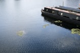 A canal boat is moored on a body of water with visible ripples and patches of algae floating on the surface. The boat is black with a reddish-brown roof and has porthole-style windows.