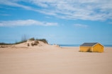 A tranquil beach scene with a bright yellow cabin situated on a sandy area. The sky is mostly clear with a few clouds, and the sea is visible in the background. There is a small sand dune with sparse vegetation to the left of the cabin.