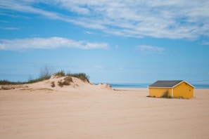 A tranquil beach scene with a bright yellow cabin situated on a sandy area. The sky is mostly clear with a few clouds, and the sea is visible in the background. There is a small sand dune with sparse vegetation to the left of the cabin.