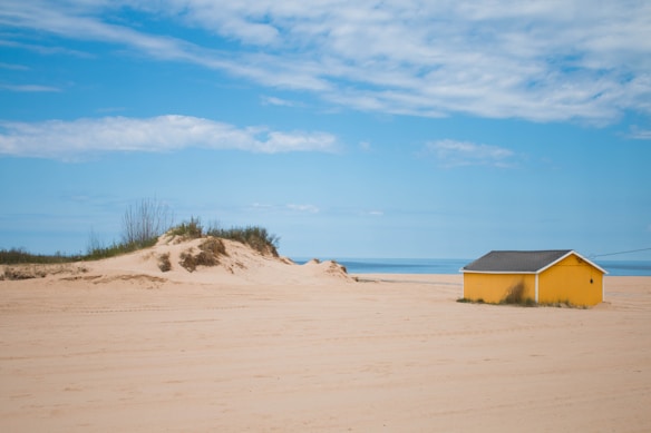 A tranquil beach scene with a bright yellow cabin situated on a sandy area. The sky is mostly clear with a few clouds, and the sea is visible in the background. There is a small sand dune with sparse vegetation to the left of the cabin.