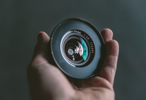 A hand is holding a large camera lens, prominently displaying the lens elements and reflections within the glass. The lens is labeled with numbers and letters, indicating specifications.