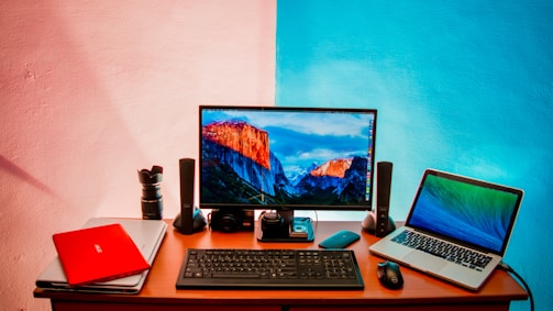 A workspace setup featuring a desk with two laptops, a desktop monitor displaying a mountain landscape, a keyboard, a mouse, speakers, a camera lens, and additional electronic accessories. The wall behind is painted in a split design with shades of pink and blue.