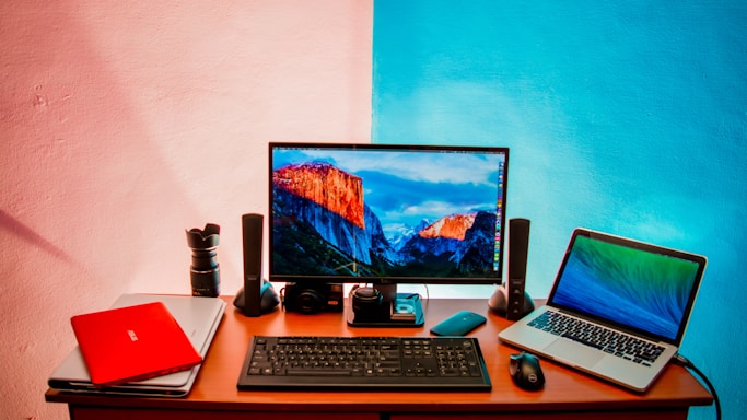 A workspace setup featuring a desk with two laptops, a desktop monitor displaying a mountain landscape, a keyboard, a mouse, speakers, a camera lens, and additional electronic accessories. The wall behind is painted in a split design with shades of pink and blue.