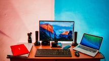 A workspace setup featuring a desk with two laptops, a desktop monitor displaying a mountain landscape, a keyboard, a mouse, speakers, a camera lens, and additional electronic accessories. The wall behind is painted in a split design with shades of pink and blue.