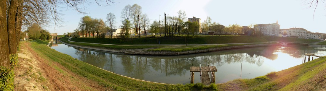 The image displays a serene riverside scene with calm water reflecting the nearby trees and sky. On either side of the river are grassy banks lined with trees, some still bare from winter. A small wooden dock extends into the river from the right bank. In the distance, buildings are visible, bathed in warm sunlight, suggesting either sunrise or sunset.