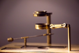 A close-up view of an antique brass microscope with multiple components, including a stage and adjustable arm mechanisms. The background is softly blurred, emphasizing the mechanical details and polished metal surfaces.