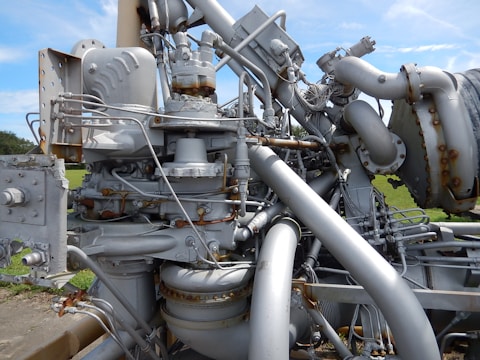 A complex and intricate assembly of industrial machinery featuring various interconnected pipes, valves, and mechanical components, with visible signs of rust and wear. The machinery is set against a backdrop of green grass and a clear blue sky.