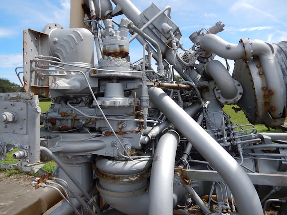 A complex and intricate assembly of industrial machinery featuring various interconnected pipes, valves, and mechanical components, with visible signs of rust and wear. The machinery is set against a backdrop of green grass and a clear blue sky.