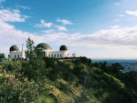 An observatory with distinctive domes sits atop a hill, surrounded by lush greenery and overlooking a sprawling cityscape in the distance. The sky is clear with scattered clouds, and a group of people is gathered near the building.