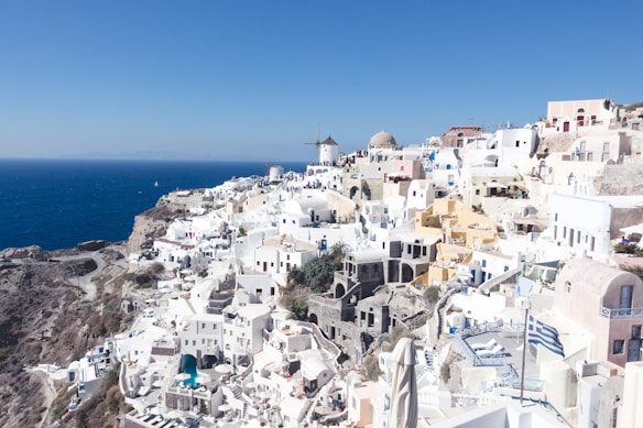A stunning coastal village with white-washed buildings sitting atop a cliff overlooking the deep blue sea. The architecture is characterized by domed roofs and narrow winding streets. A windmill and a Greek flag add to the scenic charm.