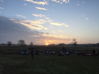 A serene outdoor setting at sunrise with several tents pitched on grassy terrain. Silhouetted figures are visible, suggesting early morning activities. The sky features a gradient of colors from dark blue to light orange and yellow as the sun peeks over the horizon. Sparse trees are scattered across the landscape, adding to the tranquil atmosphere.