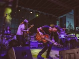 A lively band performance on stage featuring multiple musicians playing electric guitars, a bass, and drums. The musician in the foreground energetically bends forward while playing a guitar, with expressive movements. The setting includes stage lighting and musical equipment such as amplifiers and a drum set. The backdrop suggests an outdoor venue with a modern building in the background.