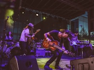 A lively band performance on stage featuring multiple musicians playing electric guitars, a bass, and drums. The musician in the foreground energetically bends forward while playing a guitar, with expressive movements. The setting includes stage lighting and musical equipment such as amplifiers and a drum set. The backdrop suggests an outdoor venue with a modern building in the background.