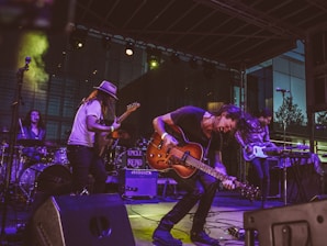 A lively band performance on stage featuring multiple musicians playing electric guitars, a bass, and drums. The musician in the foreground energetically bends forward while playing a guitar, with expressive movements. The setting includes stage lighting and musical equipment such as amplifiers and a drum set. The backdrop suggests an outdoor venue with a modern building in the background.