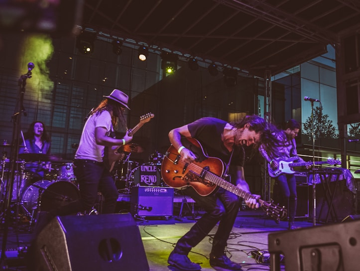A lively band performance on stage featuring multiple musicians playing electric guitars, a bass, and drums. The musician in the foreground energetically bends forward while playing a guitar, with expressive movements. The setting includes stage lighting and musical equipment such as amplifiers and a drum set. The backdrop suggests an outdoor venue with a modern building in the background.