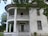 A large, old, two-story house with a white exterior and a prominent curved balcony supported by pillars. The building appears weathered and slightly neglected with peeling paint and broken windows. Surrounding the house are leafy trees, adding to the abandoned and eerie atmosphere.