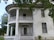 A large, old, two-story house with a white exterior and a prominent curved balcony supported by pillars. The building appears weathered and slightly neglected with peeling paint and broken windows. Surrounding the house are leafy trees, adding to the abandoned and eerie atmosphere.