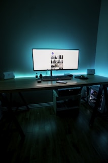 A modern workspace setup with a wide monitor displaying a web page, surrounded by ambient teal lighting. The desk is organized with a keyboard, mouse, and several electronic devices. The environment is dimly lit, creating a focused and sleek atmosphere.