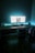 A modern workspace setup with a wide monitor displaying a web page, surrounded by ambient teal lighting. The desk is organized with a keyboard, mouse, and several electronic devices. The environment is dimly lit, creating a focused and sleek atmosphere.