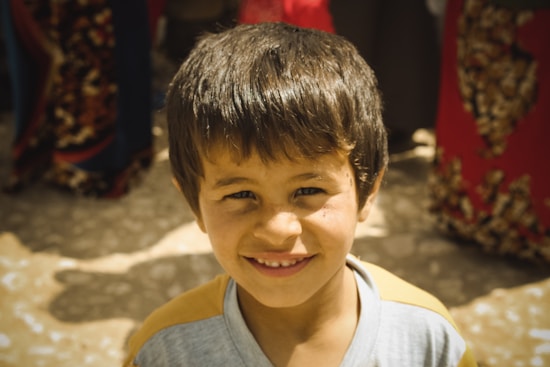 A young boy smiles warmly at the camera, standing in a sunny outdoor setting. The background contains people with colorful garments, predominantly in shades of red and blue, which are out of focus.