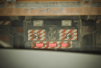 A close-up view of the back of a large vehicle, possibly a truck, with a set of red LED lights and a decorative panel featuring stylized eyes with orange and white diagonal stripes. The vehicle appears dusty, suggesting it has been used in a construction or industrial environment.
