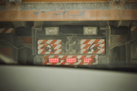 A close-up view of the back of a large vehicle, possibly a truck, with a set of red LED lights and a decorative panel featuring stylized eyes with orange and white diagonal stripes. The vehicle appears dusty, suggesting it has been used in a construction or industrial environment.
