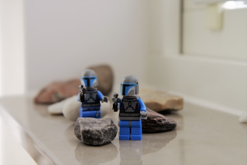Two small toy figures, resembling Star Wars Mandalorian characters, are placed on a surface scattered with smooth stones. The figures are detailed with blue and black coloring and appear to be holding small blasters. The background is out of focus, bringing attention to the figures and rocks in the foreground.
