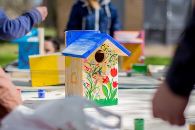 A hand-painted decorative birdhouse stands prominently on a table, featuring vibrant floral designs and a bright blue roof. Surrounding the birdhouse are various art supplies, such as paintbrushes and open jars of paint. In the background, several people engage in painting activities, positioned around the table in an outdoor setting.