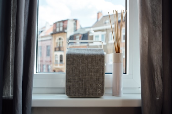 A stylish urbanecho smart speaker resting on a minimalist wooden shelf against a city apartment window backdrop.