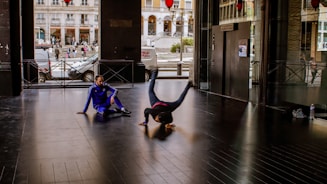Two individuals are engaged in breakdancing inside an urban building. One person is performing a headstand while the other, dressed in a blue tracksuit, sits on the floor watching. The background shows part of a street scene through glass walls, with people and vehicles visible outside.