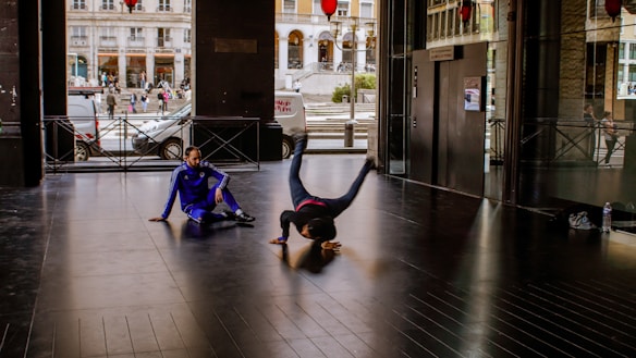 Two individuals are engaged in breakdancing inside an urban building. One person is performing a headstand while the other, dressed in a blue tracksuit, sits on the floor watching. The background shows part of a street scene through glass walls, with people and vehicles visible outside.