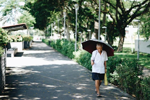 A caregiver assisting an elderly man with a morning walk in a sunny park, surrounded by green trees.