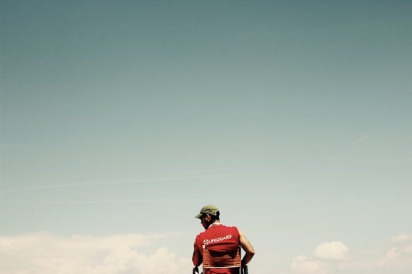 A lifeguard wearing a red vest stands against a vast, clear sky. The vest is prominently labeled with the word 'Lifeguard', and the lifeguard appears to be looking out into the distance. The background is composed of pale blue and white hues, suggesting an open and calm environment.
