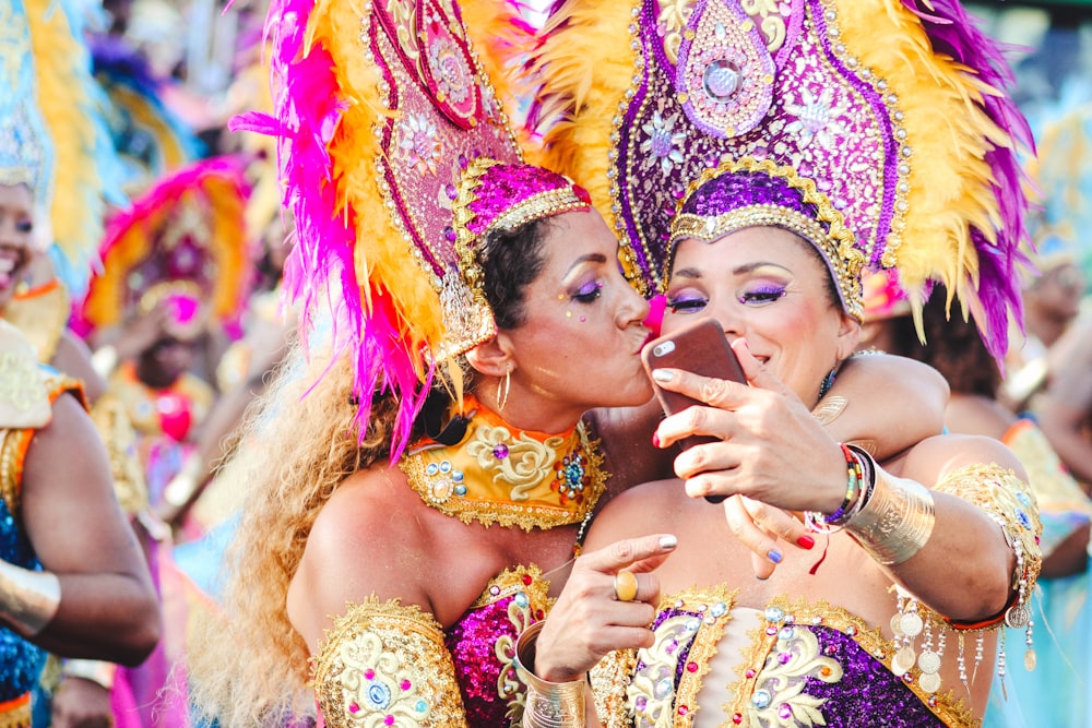 두 명의 여성 카니발 댄서가 셀카를 위해 포즈를 취하고 한 명이 다른 사람의 뺨에 키스합니다.