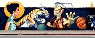 A vibrant mural featuring a woman in traditional dress with a red eye mask, a tiger emerging from water, and a warrior figure with an ornate headpiece and a staff. The scene includes two large planets, one resembling Saturn, set against a dark background.