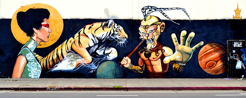 A vibrant mural featuring a woman in traditional dress with a red eye mask, a tiger emerging from water, and a warrior figure with an ornate headpiece and a staff. The scene includes two large planets, one resembling Saturn, set against a dark background.