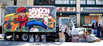 A delivery truck with vibrant graffiti art depicting a cartoon character and the words 'Dragon School'. The truck is parked on a street lined with shops, featuring signs in both English and Chinese. Numerous cardboard boxes are piled up near the truck, possibly for loading or unloading. Several people are visible in the background, engaging in activities.
