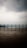 A wooden deck, glistening with moisture, stretches across the foreground with a simple railing separating it from the expansive view of a calm body of water. In the background, mountains shrouded in mist rise under a sky thick with overcast clouds.