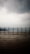 A wooden deck, glistening with moisture, stretches across the foreground with a simple railing separating it from the expansive view of a calm body of water. In the background, mountains shrouded in mist rise under a sky thick with overcast clouds.