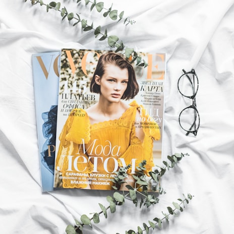 Two magazine covers are placed side by side on a white cloth, with one prominently featuring a person in a yellow outfit. Nearby, there is a pair of black-rimmed eyeglasses and a few sprigs of eucalyptus leaves, adding a natural touch to the composition.