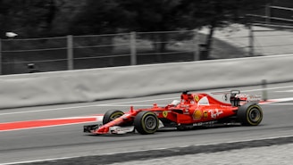 A red Formula 1 race car speeds along a track. The car is adorned with various logos and sponsors, featuring sleek aerodynamic design elements. The background is a blurred mix of gray and black, emphasizing the motion and speed of the vehicle.